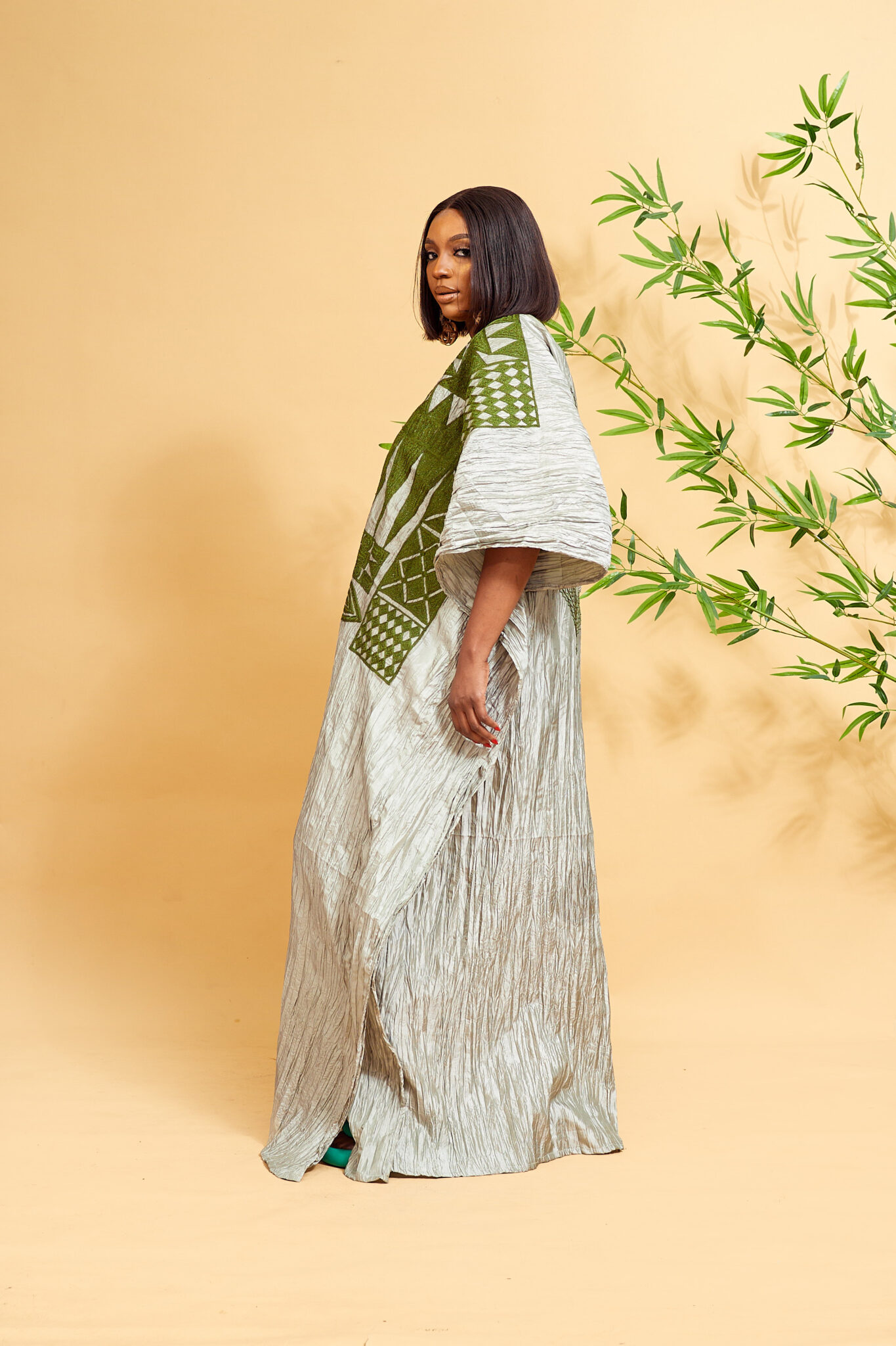 Lady wearing a light green khaftan with dark green embroidery. She is standing in a studio with a light yellow background and a green bamboo shoot